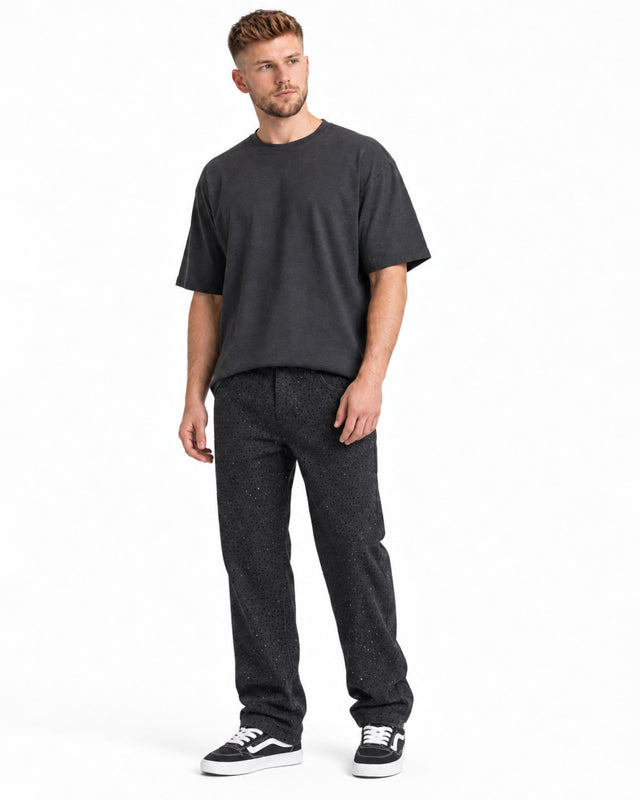 A man stands wearing a loose-fitting dark t-shirt and patterned black pants, paired with black and white sneakers.