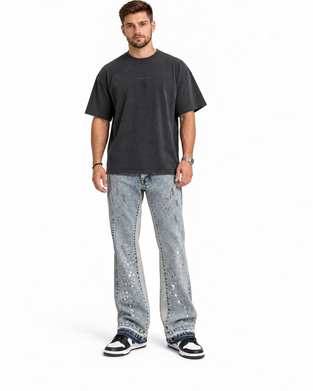 A man stands wearing a dark oversized t-shirt, distressed light jeans, and black and white sneakers.