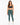 A woman stands confidently in a dark sports bra and teal leggings, paired with white sneakers.