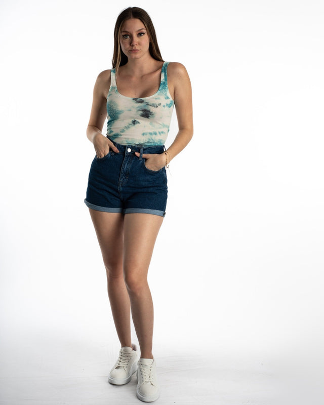 A model poses in a tie-dye tank top and high-waisted denim shorts, complemented by white sneakers.