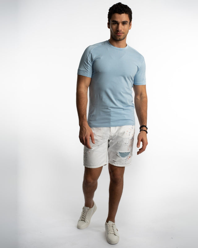 A man stands confidently in a light blue short-sleeve shirt and distressed white shorts, paired with casual sneakers.