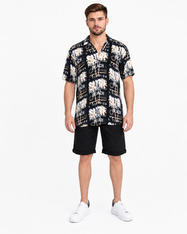 A man stands wearing a short-sleeve button shirt with a black and white graphic print, paired with black shorts.