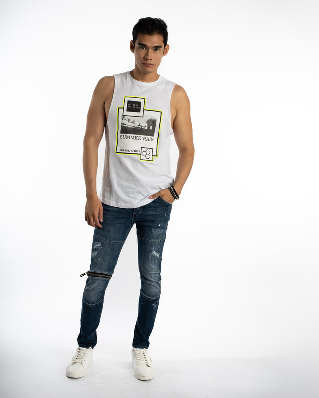 A young man poses in a sleeveless white top with a graphic design, paired with distressed blue jeans and white sneakers.