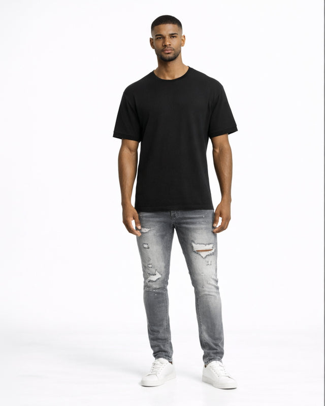 A man stands confidently in a black t-shirt, distressed gray jeans, and white sneakers against a plain background.