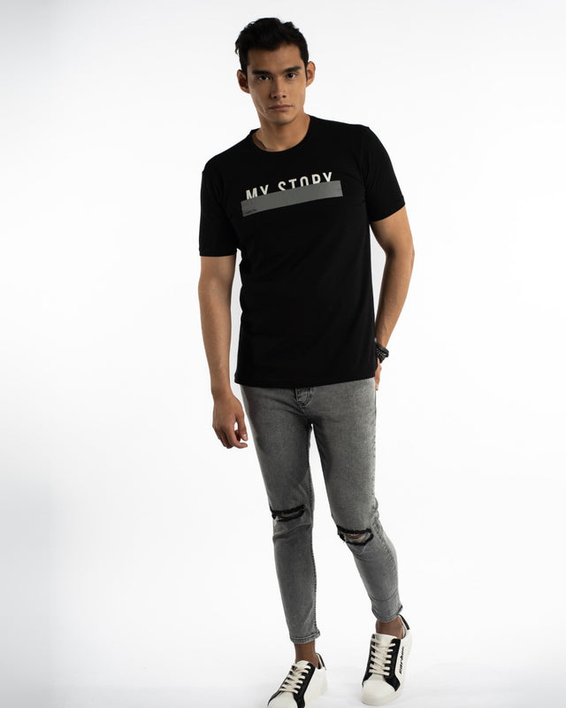 A model wearing a black t-shirt with a graphic design, paired with distressed gray jeans and stylish sneakers.