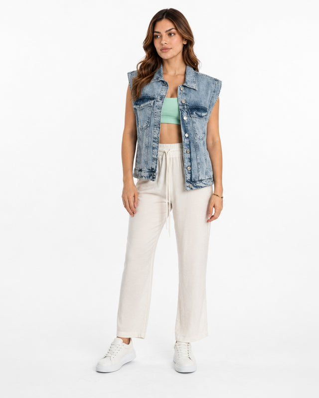 A woman stands confidently in a sleeveless denim vest over a green crop top, paired with light pants and white sneakers.