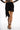 Model wearing Harlow Black Mini Skirt featuring a draped wrap front, asymmetrical hem, and black fringe tassel trim.