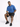 A man sits on a white pedestal wearing a blue oversized t-shirt and black shorts, with black sneakers.