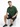 A man sits on a white cube wearing a green oversized t-shirt, black shorts, and black sneakers with white stripes.