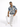 A man sits on a white pedestal, wearing a short-sleeve, blue and beige patterned shirt with white ripped jeans.