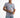 Man in lilac streetwear graphic tee with a teal chest logo and light wash distressed jeans on white studio background.