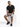 A man sits on a white pedestal wearing a black t-shirt, gray shorts, and black sneakers, showcasing a casual style.