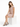 A woman sits on a white pedestal wearing a soft pink dress and black strappy heels, exuding a relaxed elegance.