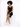 A woman with curly hair sits on a white cube, wearing a fitted black dress and white sneakers.