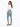A woman with long hair wears a white long-sleeve top and light blue jeans, posing sideways in casual footwear.
