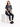 A woman sits on a white cube, wearing a graphic tee, dark jeans, and black sneakers, with a relaxed expression.