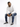 A man sits on a white pedestal wearing a light hoodie, distressed jeans, and black sneakers, looking thoughtfully to...
