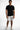 A man stands confidently in a fitted black t-shirt and distressed white shorts, paired with casual sneakers.