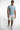 A man stands confidently in a light blue short-sleeve shirt and distressed white shorts, paired with casual sneakers.