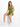 A model sits on a white cube wearing a fitted olive green one-shoulder top and matching shorts with white sneakers.