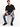 A man sits on a white pedestal wearing a loose black t-shirt, distressed blue jeans, and black sneakers.