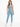 A woman stands confidently in a light blue one-shoulder crop top and matching leggings, paired with white sneakers.