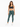 A woman stands confidently in fitted green leggings and a dark sports bra, paired with white sneakers.