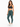 A woman stands confidently in fitted teal leggings and a dark sports bra, paired with white sneakers.
