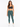 A woman stands confidently in a dark sports bra and teal leggings, paired with white sneakers.
