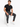 A man sits on a white cube, wearing a fitted black t-shirt and dark gray pants with white sneakers.