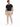 A man stands wearing a black t-shirt and beige evening pants, paired with white sneakers and a chain detail.