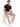 A man sits on a white cube wearing beige evening pants, a black t-shirt, and white sneakers.
