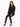 A model wearing a sheer black button-up shirt and fitted black pants with side slits, seated on a stool.