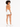A model with curly hair wearing an orange bodysuit and white shorts, standing with her back to the camera.