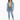 A woman stands wearing a cropped denim jacket over a light green top, paired with high-waisted jeans and white sneakers.