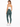 A woman in a fitted white crop top and green leggings poses from the back, showcasing a casual athletic look.