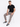 A man sits on a white stool wearing a black t-shirt, gray joggers, and black and white sneakers.