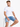 A man sits on a stool wearing a plain white t-shirt and denim shorts, with a neutral background.
