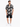 A man wearing a short-sleeve, patterned shirt with a Miami Beach theme, paired with black shorts and white sneakers.