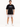 A man stands wearing a black oversized t-shirt and light gray rolled-up shorts, paired with white sneakers.