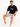 A man sits on a white cube wearing a black oversized t-shirt and light gray shorts, paired with white sneakers.