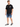 A man stands wearing a black t-shirt, denim shorts with rolled cuffs, and black sneakers.