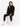 A model sits on a white cube wearing black sweatpants and a matching top, paired with white high-top sneakers.