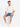 A man sits on a white cube wearing a t-shirt and denim shorts, paired with white sneakers.