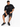 A man sits on a white cube wearing a black graphic t-shirt, distressed black shorts, and black sneakers.