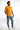 A man stands confidently in a fitted orange shirt and distressed blue jeans, paired with white sneakers.