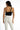 A woman stands with her back to the camera, wearing a black one-shoulder top and white fitted pants.