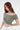 A model poses in a soft, off-the-shoulder, cropped top in a muted green shade, paired with white pants.