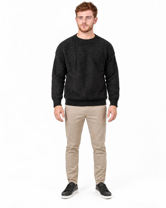 A man stands in a black textured sweatshirt, beige pants, and black sneakers against a white background.
