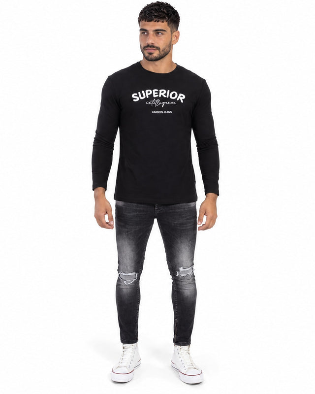A man stands wearing a long-sleeve black shirt with white text, paired with distressed gray jeans and white sneakers.